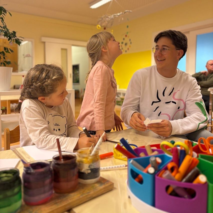 Ein Klassenzimmer mit einem Lehrer und zwei Mädchen, eines steht und schaut nach oben, das andere sitzt an einem Schreibtisch mit Malutensilien, der Lehrer lächelt und hält Papier. Ein Mann beobachtet aus der Ferne.