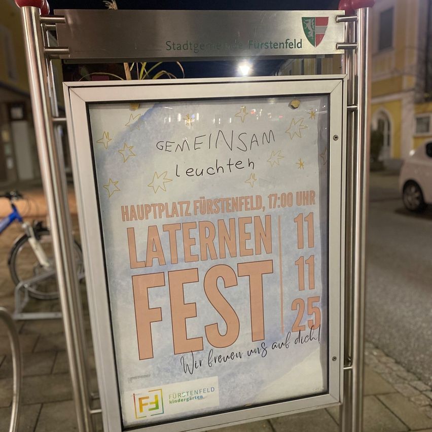 Ein Schild zeigt 'Gemeinsam leuchten wir', 'Laternenumzug in Furstenfeld, 17:00 Uhr' und 'Laternen 11 11 25'. Es steht auf einem Metallständer vor einem Gebäude.