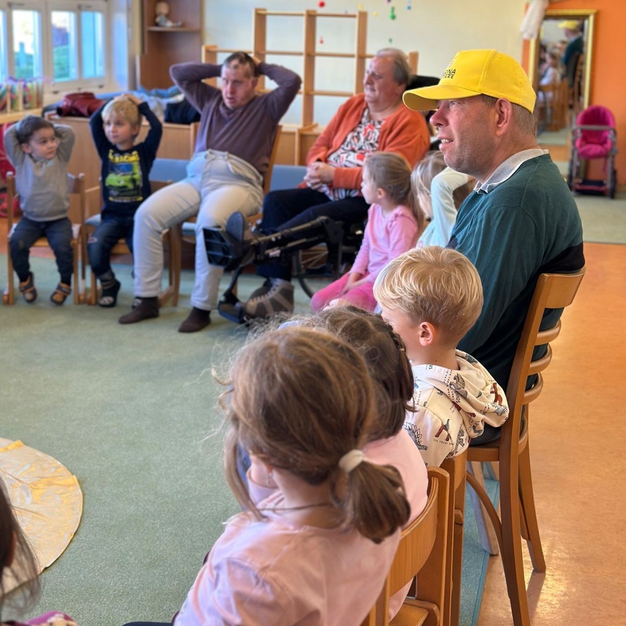Eine Gruppe von Erwachsenen und Kindern sitzt im Kreis. Ein Mann mit gelber Kappe sitzt in der Mitte. Kinder sitzen auf Stühlen, während Erwachsene auf Bänken sitzen. Ein Kinderwagen steht in der Nähe.