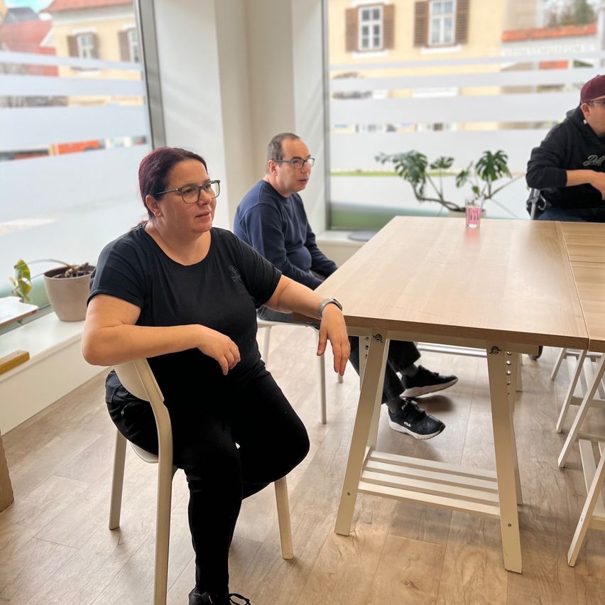 Drei Personen sitzen in einem modernen Büro. Eine Frau mit Brille und schwarzer Kleidung lehnt sich an einen Tisch. Ein Mann in einem blauen Shirt sitzt neben ihr, und ein anderer Mann im Rollstuhl trägt eine Kappe. Topfpflanzen und ein Gebäude sind durch die Fenster sichtbar.
