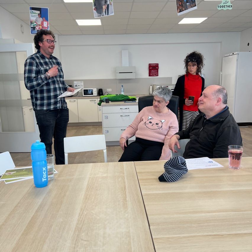 Ein Mann steht und spricht, ein Papier in der Hand. Eine Frau sitzt neben einem Mann, beide lächeln. Eine Frau in Rot steht hinter ihnen. Ein Tisch mit einer Flasche, einem Becher und einem Hut. Ein Kühlschrank, eine Mikrowelle und Poster schmücken den Hintergrund.