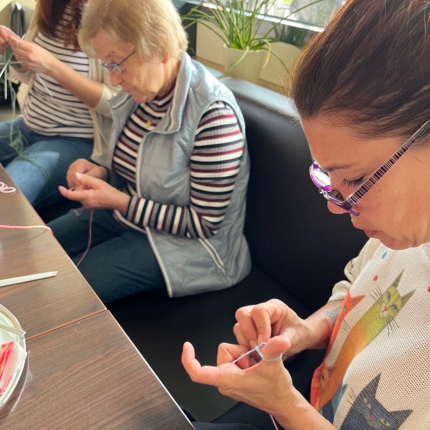 Three women sit together at a table, engrossed in their knitting. One woman wears glasses, while another has a colorful cat-printed shirt.