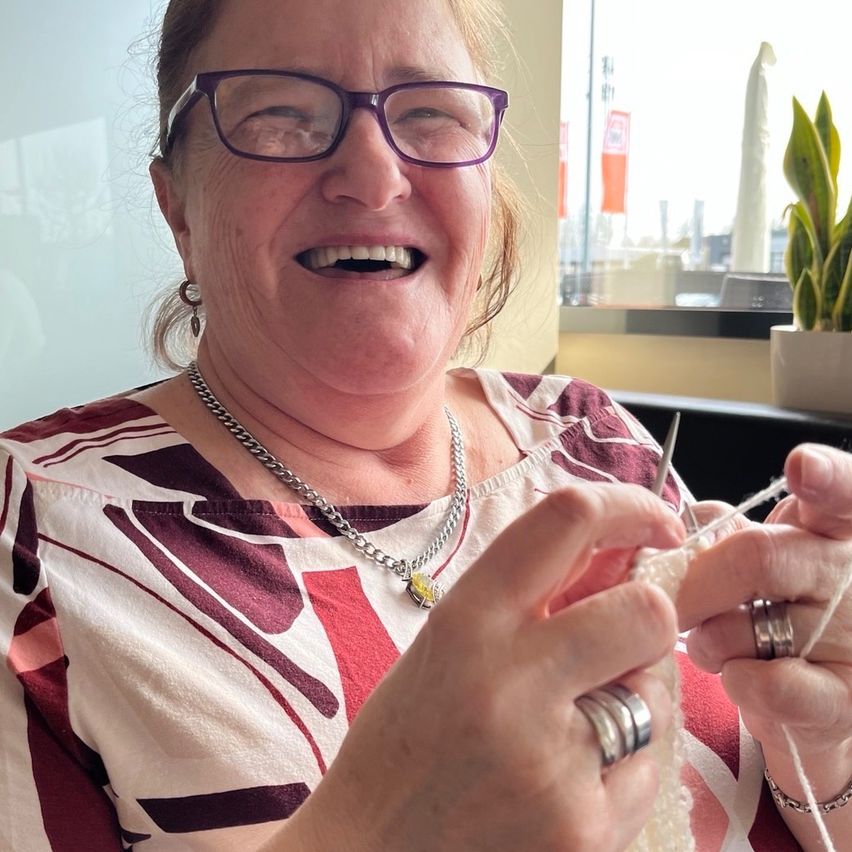 A smiling woman with glasses holds knitting needles and yarn. She wears a necklace, rings, and a patterned shirt. A potted plant is behind her.
