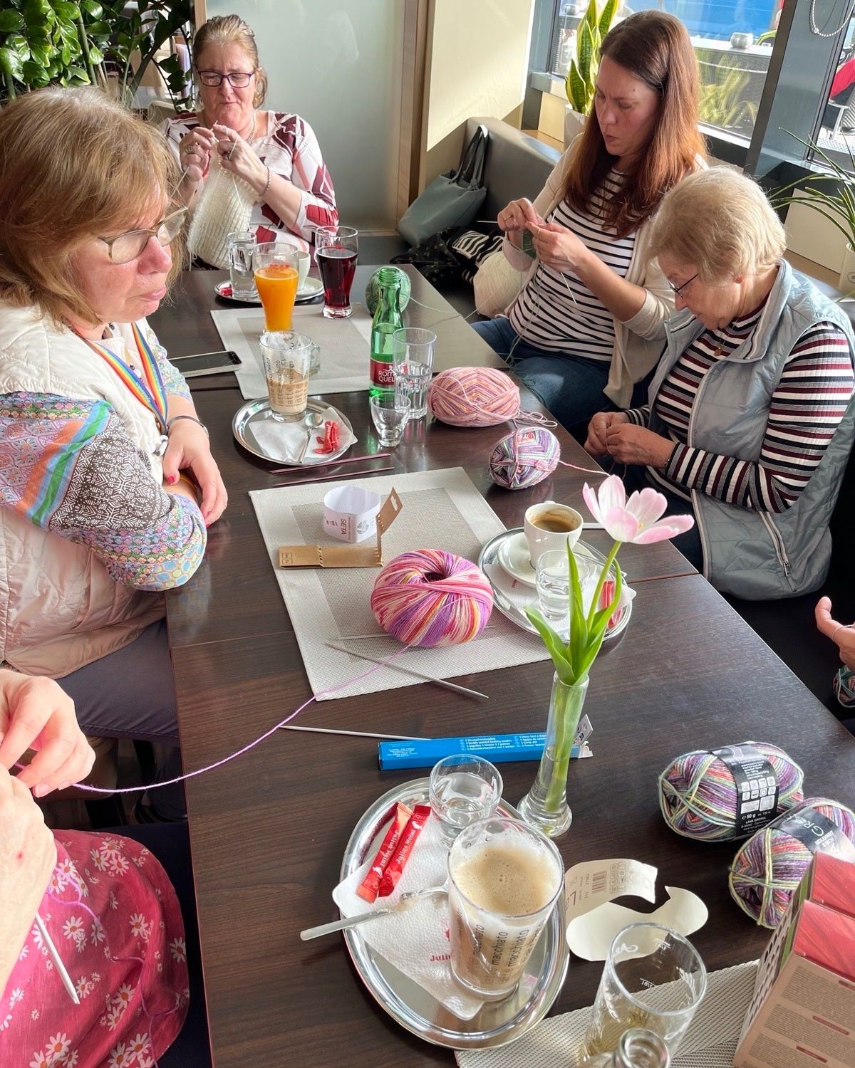 Several women sit around a table, knitting. A table has a cup of coffee, a saucer, a flower, and yarn. The women wear glasses and jackets. Glassware and drinks are on the table.