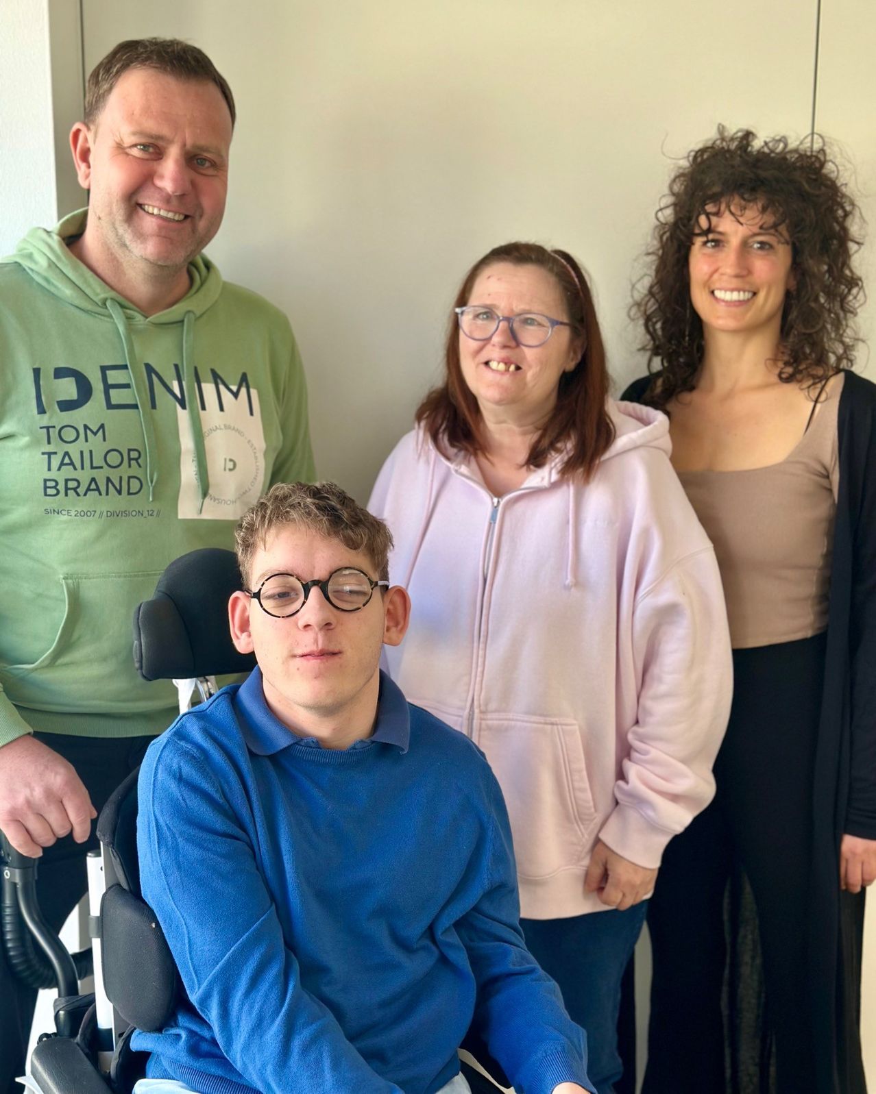 Four people are standing in a room. One is in a wheelchair. The man on the left wears a green hoodie with the words 'ID DENIM'. The woman next to him has glasses. The other two women have curly hair.