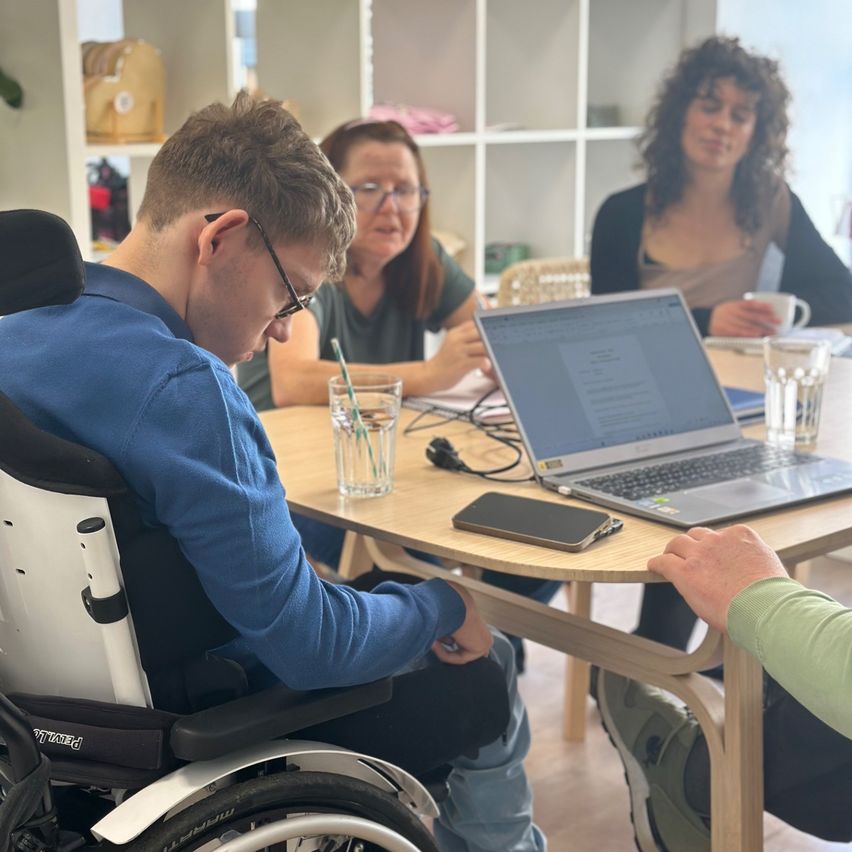 A man in a wheelchair is engaged in a discussion with two women. They are seated around a wooden table, with a laptop, a phone, and a glass of water on the table.