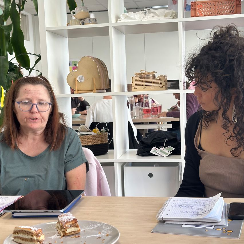 Two women sit at a desk, one with food on a plate, the other with a notebook and phone. A white shelf with various items is behind them.