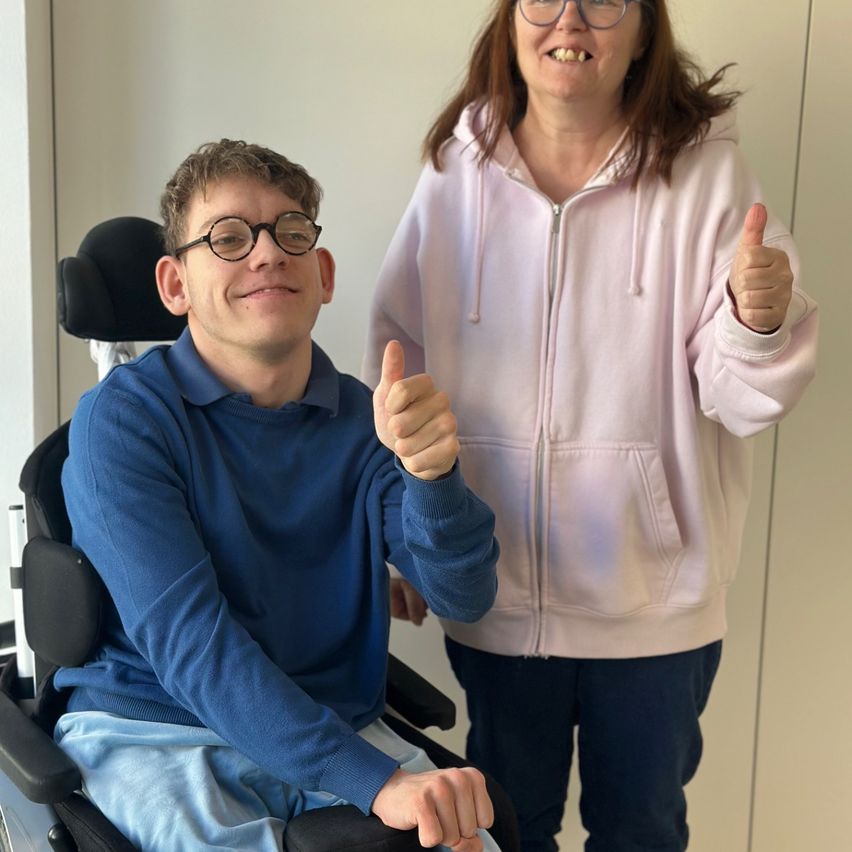 A man in a wheelchair and a woman in a pink hoodie are giving thumbs up in a room.