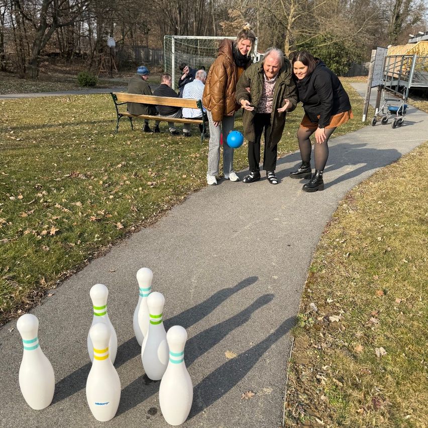 Four women are playing a game with bowling pins on a path in a park. A soccer goal and a bench are in the background.