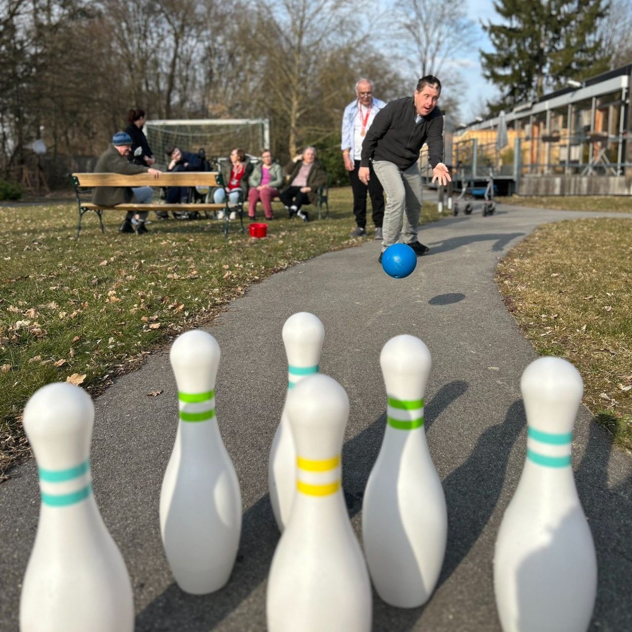 A man is playing with a blue ball on a pathway with bowling pins, while people sit on benches in the background.