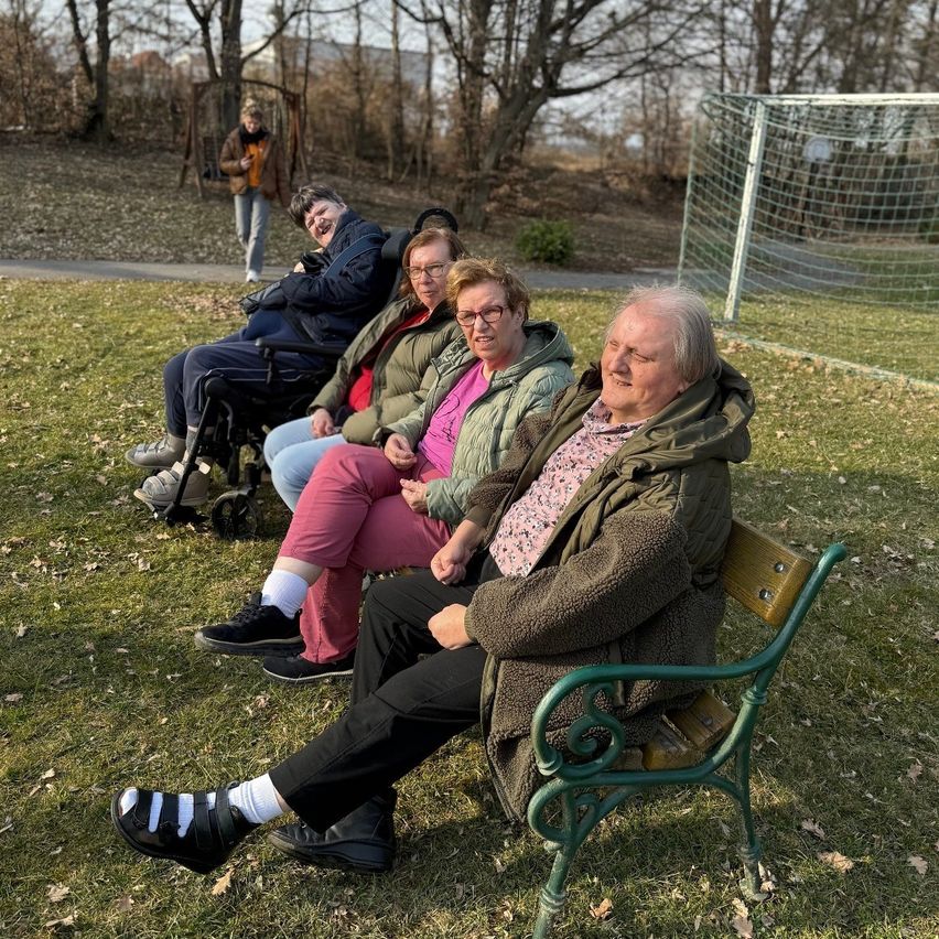 A group of older adults are sitting on a bench in a park. They are smiling and appear to be enjoying each other's company. Behind them is a soccer goal.