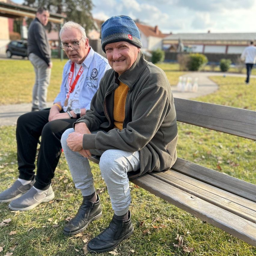 Two men are sitting on a bench in a park. One is wearing a blue hat, the other has a medal around his neck. Behind them, a man stands near a building.