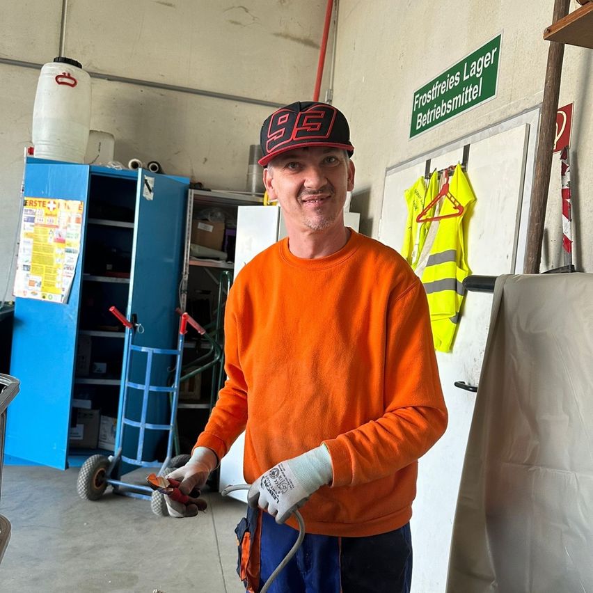 Ein Mann in einem orangefarbenen Sweatshirt steht in einer Garage und hält eine Zange in der Hand. Hinter ihm befindet sich ein blauer Lagerschrank, ein großer weißer Behälter und ein Schild mit der Aufschrift 'Frostfreies Lager Betriebsmittel'.