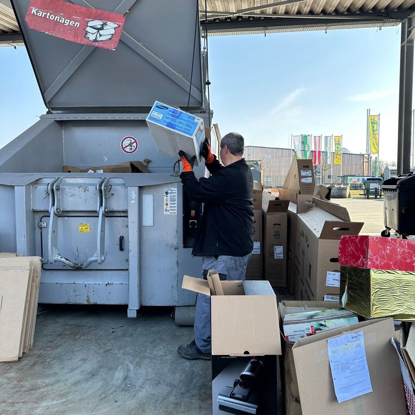 Ein Mann in Handschuhen leert eine Schachtel in einen Recyclingbehälter mit einem Schild, das 'Kartonagen' besagt. Viele weitere Kartons sind in der Nähe gestapelt, und ein Banner hängt über dem Behälter.