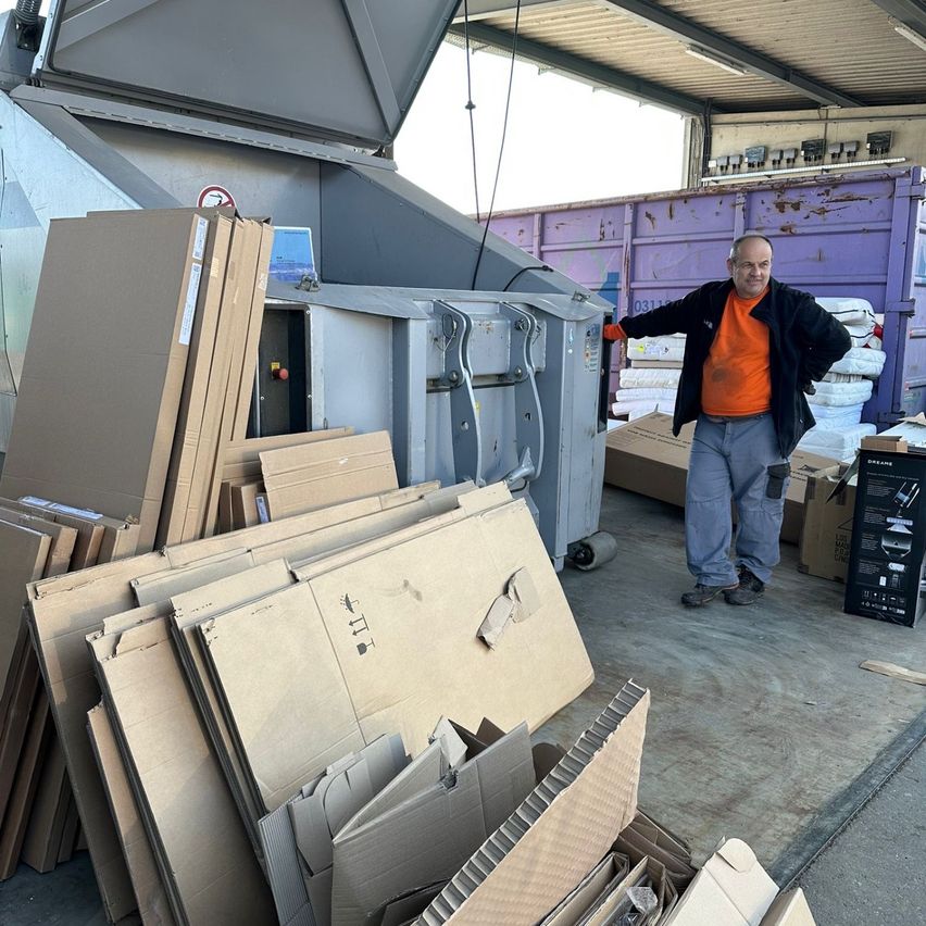 Ein Mann steht neben einem großen Haufen Pappkartons und schaut zur Kamera. Hinter ihm befindet sich eine große graue Recyclingmaschine mit offener Tür. Im Hintergrund sind auch einige Matratzen und andere Gegenstände zu sehen.