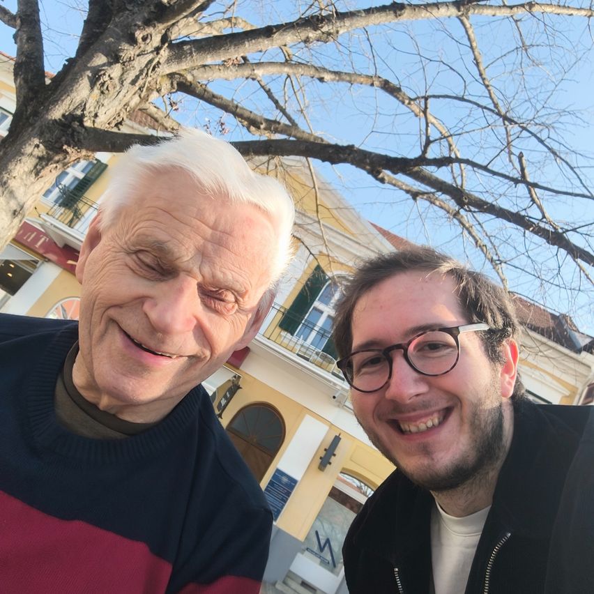 Zwei Männer stehen unter einem Baum vor einem Gebäude mit gelben Wänden und grünen Fensterläden. Der ältere Mann hat weiße Haare und trägt einen Pullover. Der jüngere Mann trägt eine Brille und eine Jacke. Sie lächeln beide.