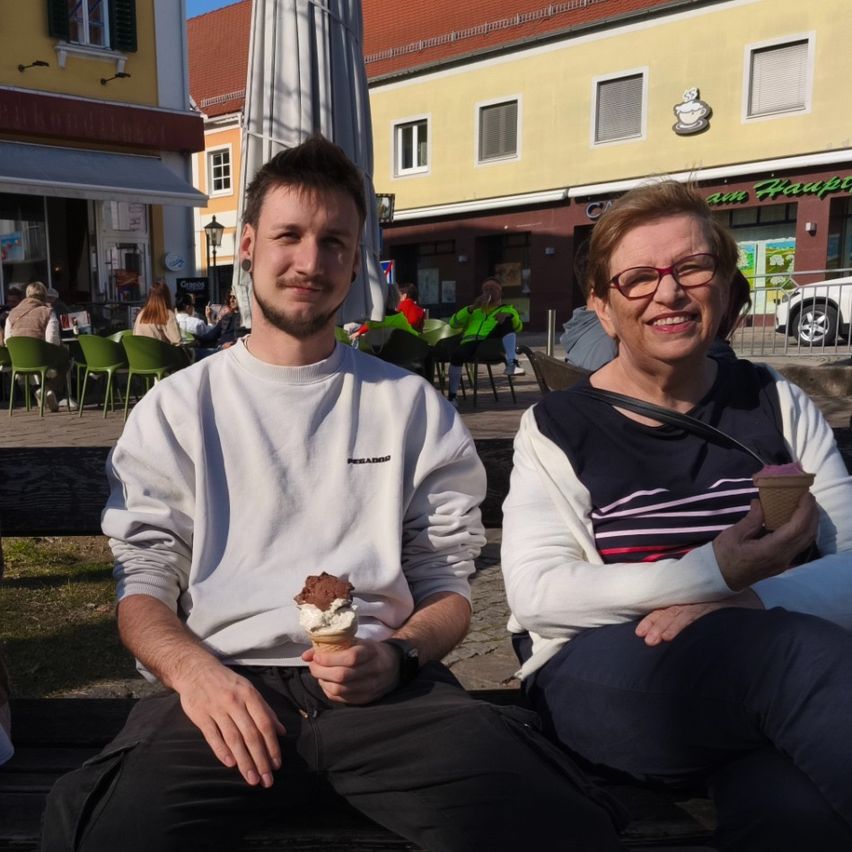 Ein Mann und eine Frau sitzen lächelnd auf einer Bank im Freien. Der Mann hält einen Schokoladeneisbecher, und die Frau hat einen rosafarbenen Eisbecher. Sie sind von Stühlen und Tischen umgeben, und ein Gebäude mit einem Café-Schild ist im Hintergrund.