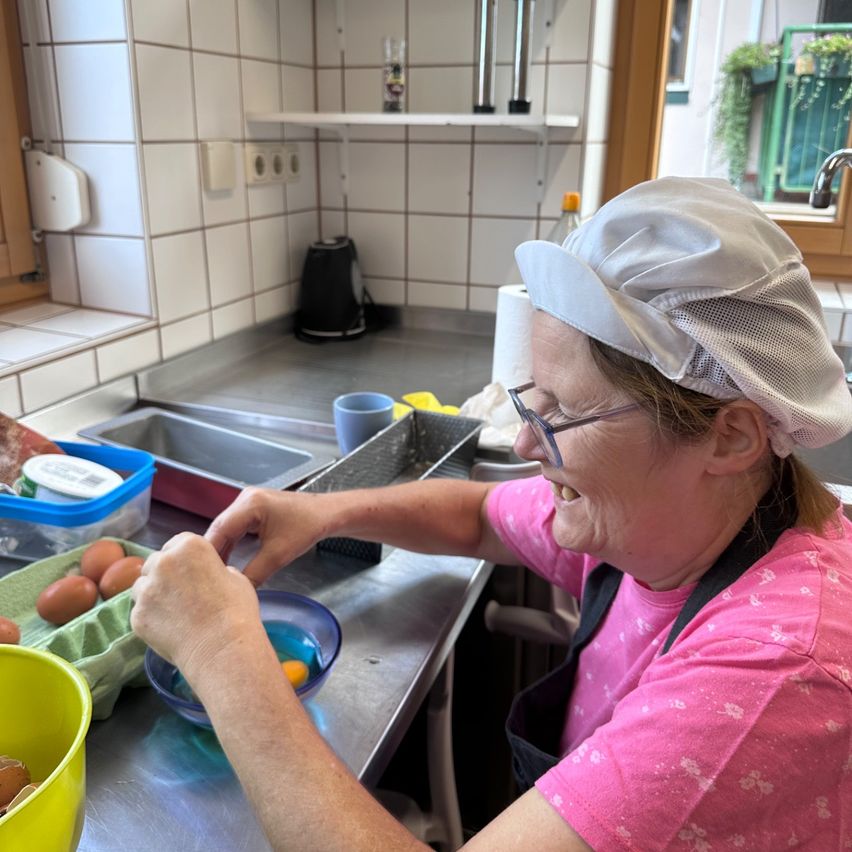A woman in a pink shirt and a white chef's hat is cracking eggs into a bowl in a kitchen.
