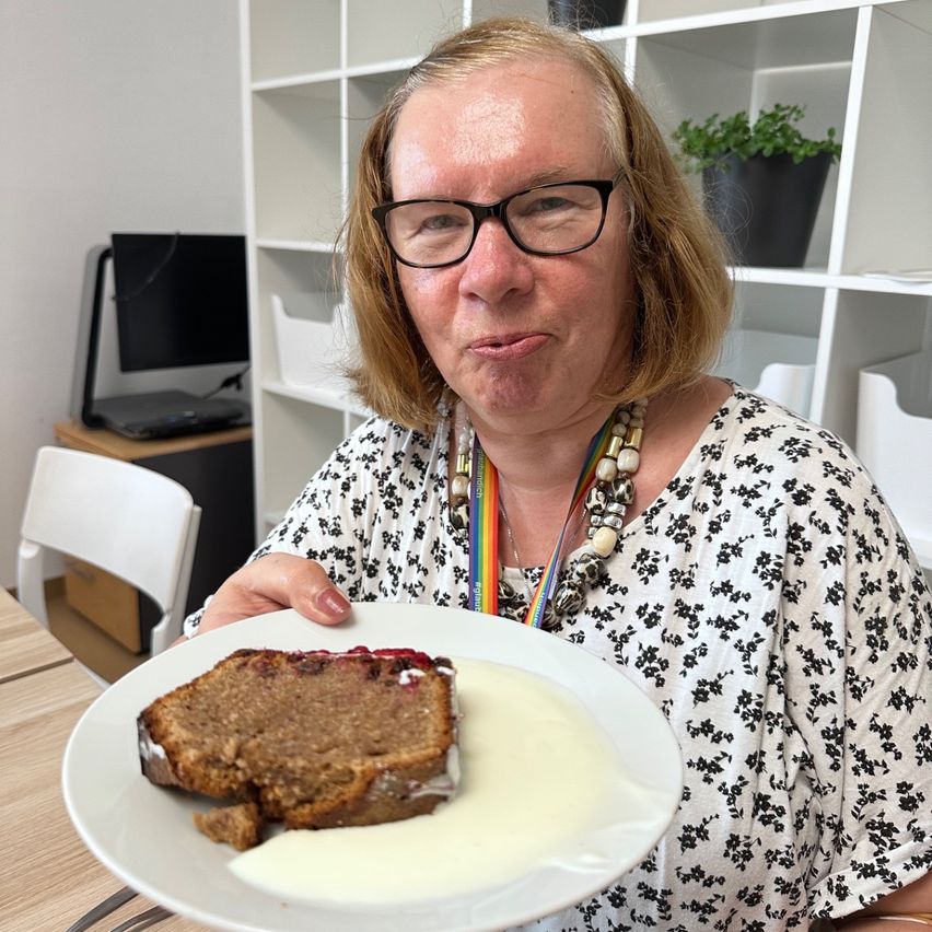 A woman with glasses and a floral shirt holds a plate with a slice of banana bread and cream. She is in a room with white shelves and a computer.