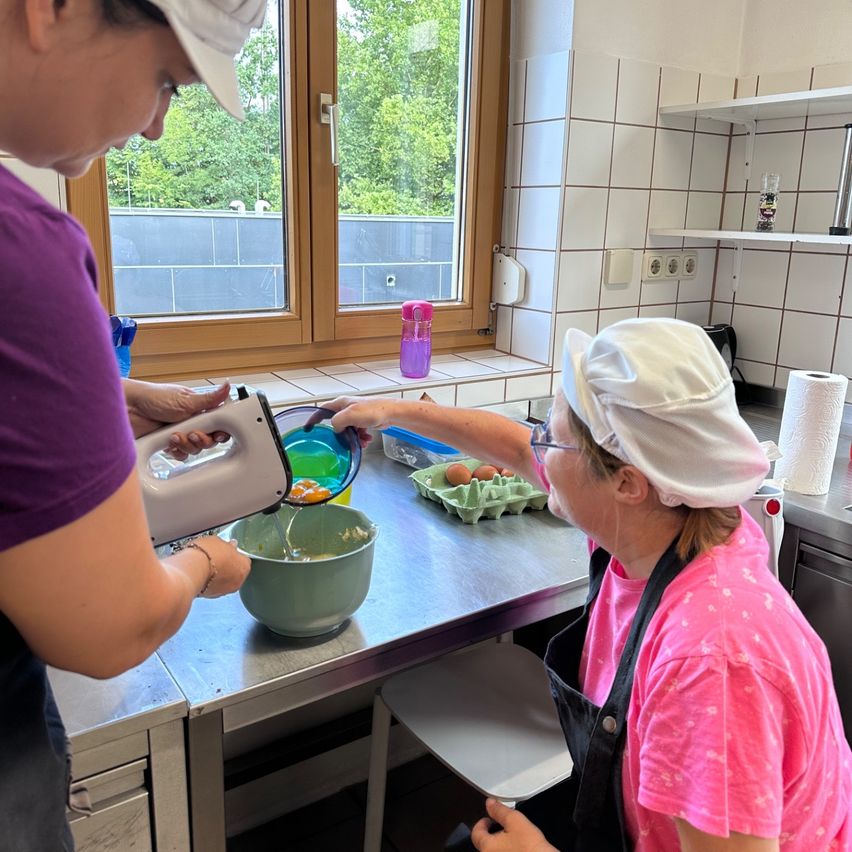 Two women in a kitchen, one holding a mixer and the other holding a bowl, both wearing aprons and caps. There is a window behind them with a view of trees.