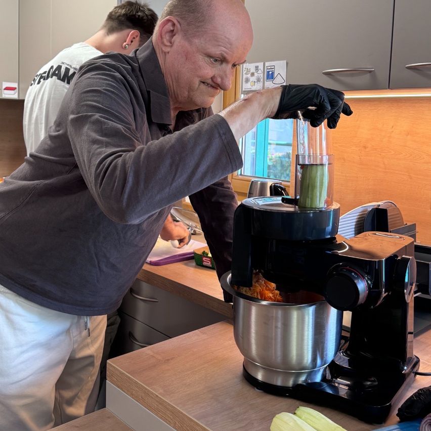 Ein älterer Mann steht in einer Küche und benutzt einen Mixer, um Zutaten zu pürieren. Er trägt Handschuhe und scheint konzentriert auf seine Aufgabe. Eine weitere Person ist im Hintergrund, möglicherweise bei der Essenszubereitung.