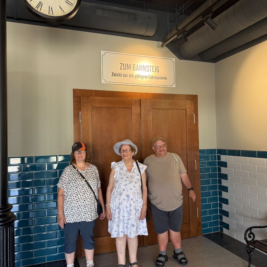 Drei Personen stehen vor einer Holztür mit einem Schild, das 'Zum Bahnsteig' besagt. Die Frau links trägt ein Blumenhemd und eine Mütze, die Frau in der Mitte trägt ein weißes Kleid und eine Brille, und der Mann rechts trägt ein beiges Hemd und Shorts. Sie befinden sich in einem Raum mit blauen Fliesen und einer Uhr darüber.