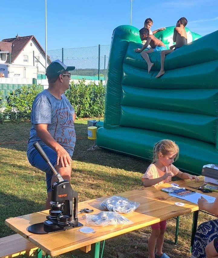 Ein Mann und ein Mädchen sitzen an einem Tisch mit verschiedenen Gegenständen, während drei Jungen auf einem großen grünen aufblasbaren Schlitten im Hintergrund spielen.
