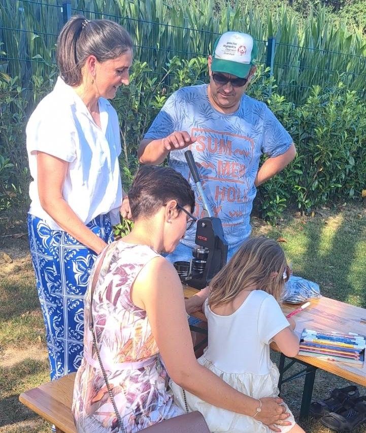 Eine Familie ist um einen Tisch im Freien versammelt, wobei ein junges Mädchen ein Mikroskop benutzt. Die Frau im Blumenkleid schaut auf das Mädchen, während der Mann im Hut beobachtet. Hinter ihnen lächelt eine Frau in einem weißen Hemd.