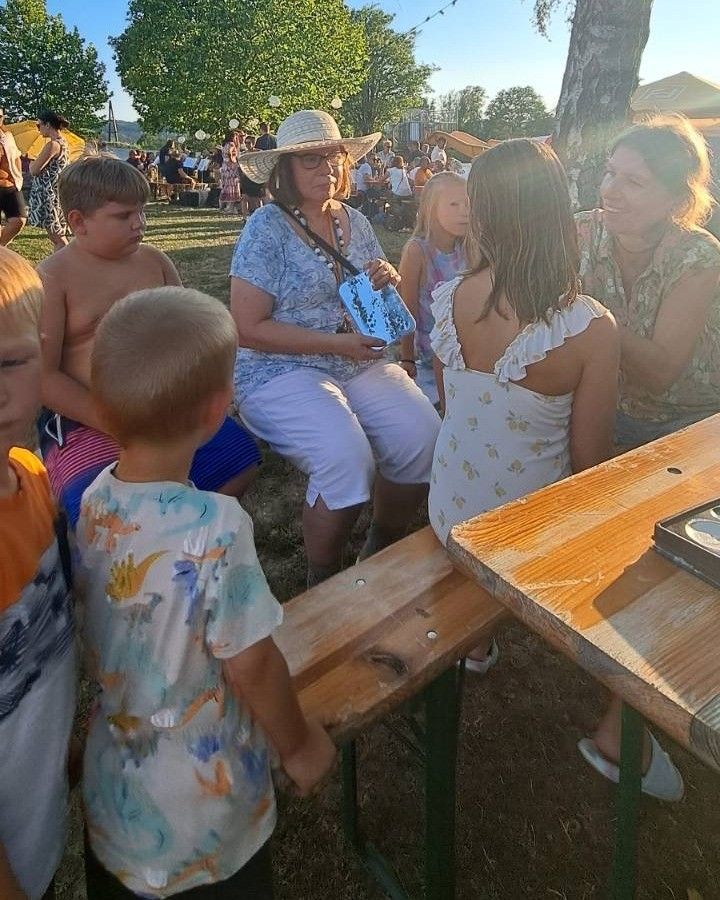 Eine Gruppe von Menschen, einschließlich Kindern und Erwachsenen, versammelt sich im Freien. Eine Frau in einem Hut hält ein blaues Objekt, und ein Mädchen in einem weißen Kleid schaut sie an. In der Nähe ist ein Holztisch mit einem Buch zu sehen.