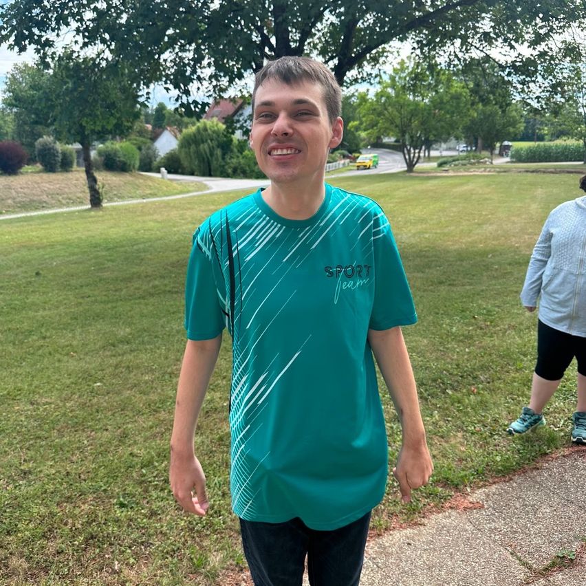 A young man with a cheerful expression stands in a park wearing a teal shirt with white stripes and the word 'SPORT' on it. In the background, a person walks on a sidewalk.