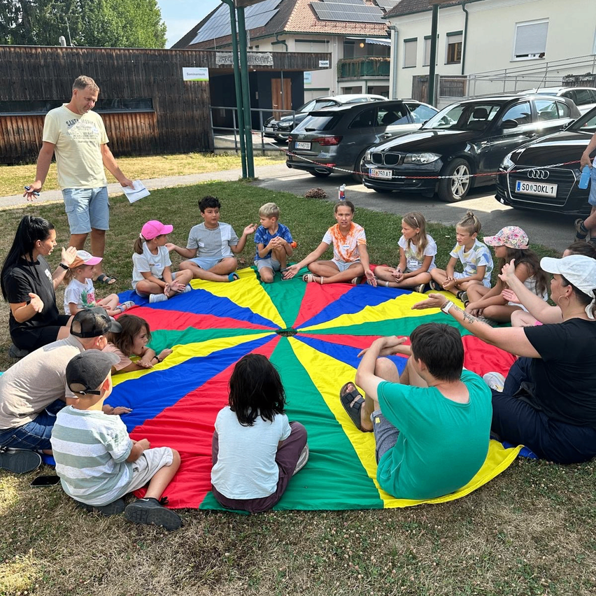 Eine Gruppe von Kindern und Erwachsenen sitzt im Kreis auf einem bunten Teppich auf dem Rasen. Ein Mann steht in der Nähe und hält ein Stück Papier. Im Hintergrund sind geparkte Autos und ein Gebäude mit Solarmodulen zu sehen.