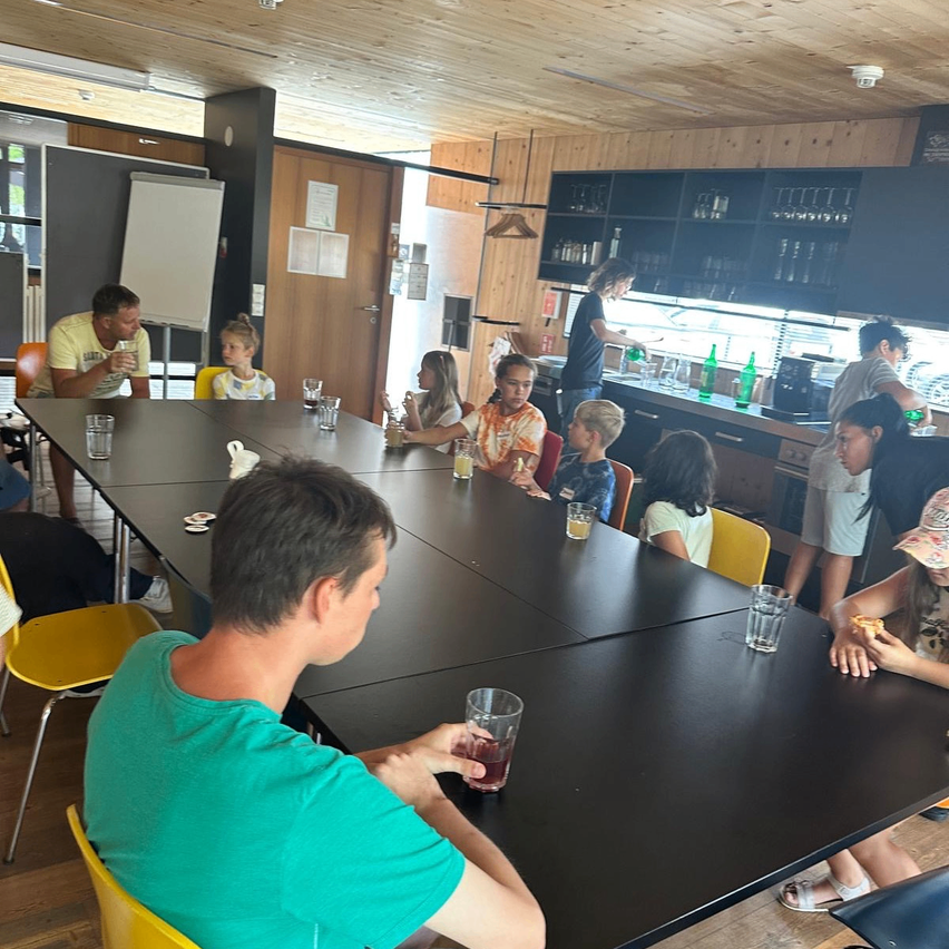 Eine Gruppe von Menschen sitzt um einen langen Tisch in einem Raum mit Holzwänden und -decke. Einige trinken aus Gläsern, während andere essen. Eine Frau steht hinter der Theke im Hintergrund.