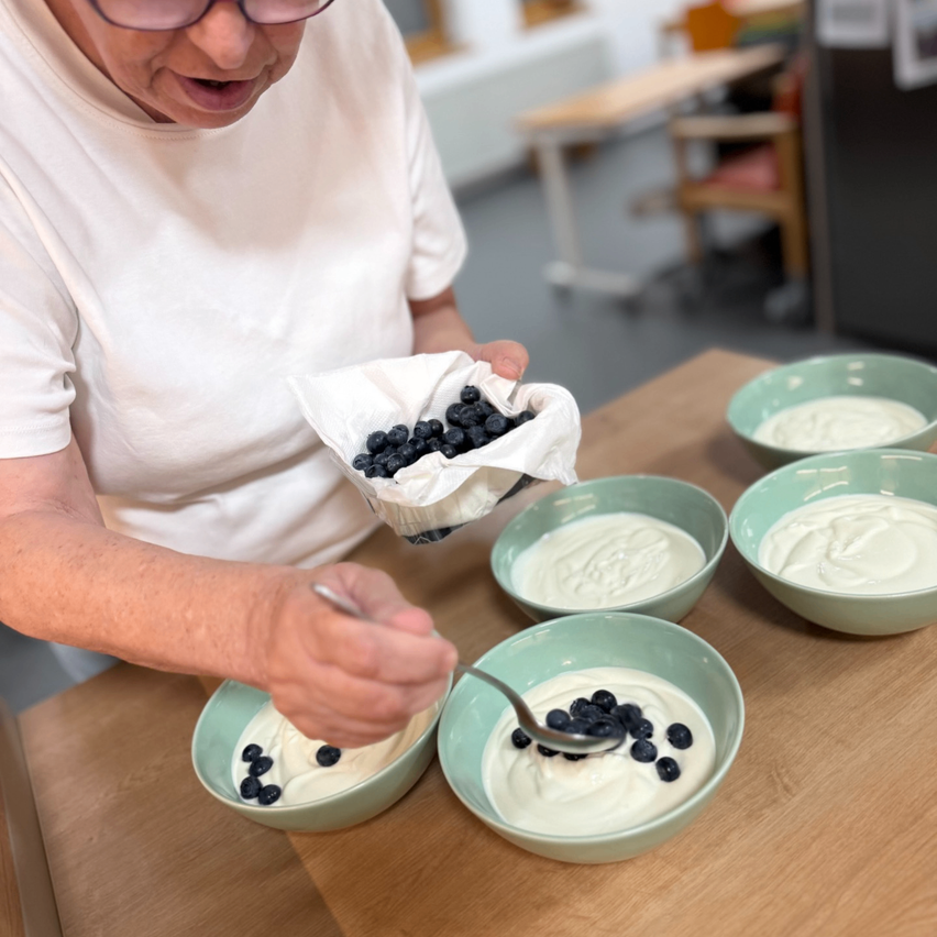 Eine Frau in einem weißen T-Shirt bereitet eine Schüssel Joghurt mit Blaubeeren zu. Sie hält einen Stoffbeutel mit Blaubeeren und ist dabei, sie dem Joghurt hinzuzufügen. Es gibt vier Schüsseln Joghurt auf dem Tisch.