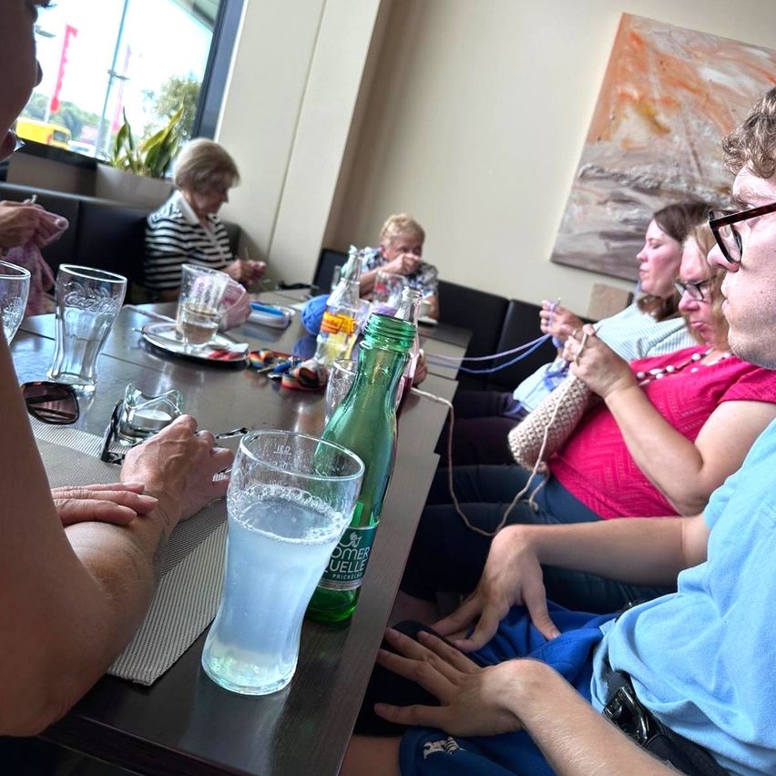 A group of people are sitting around a table, knitting. One person is holding a glass of water.