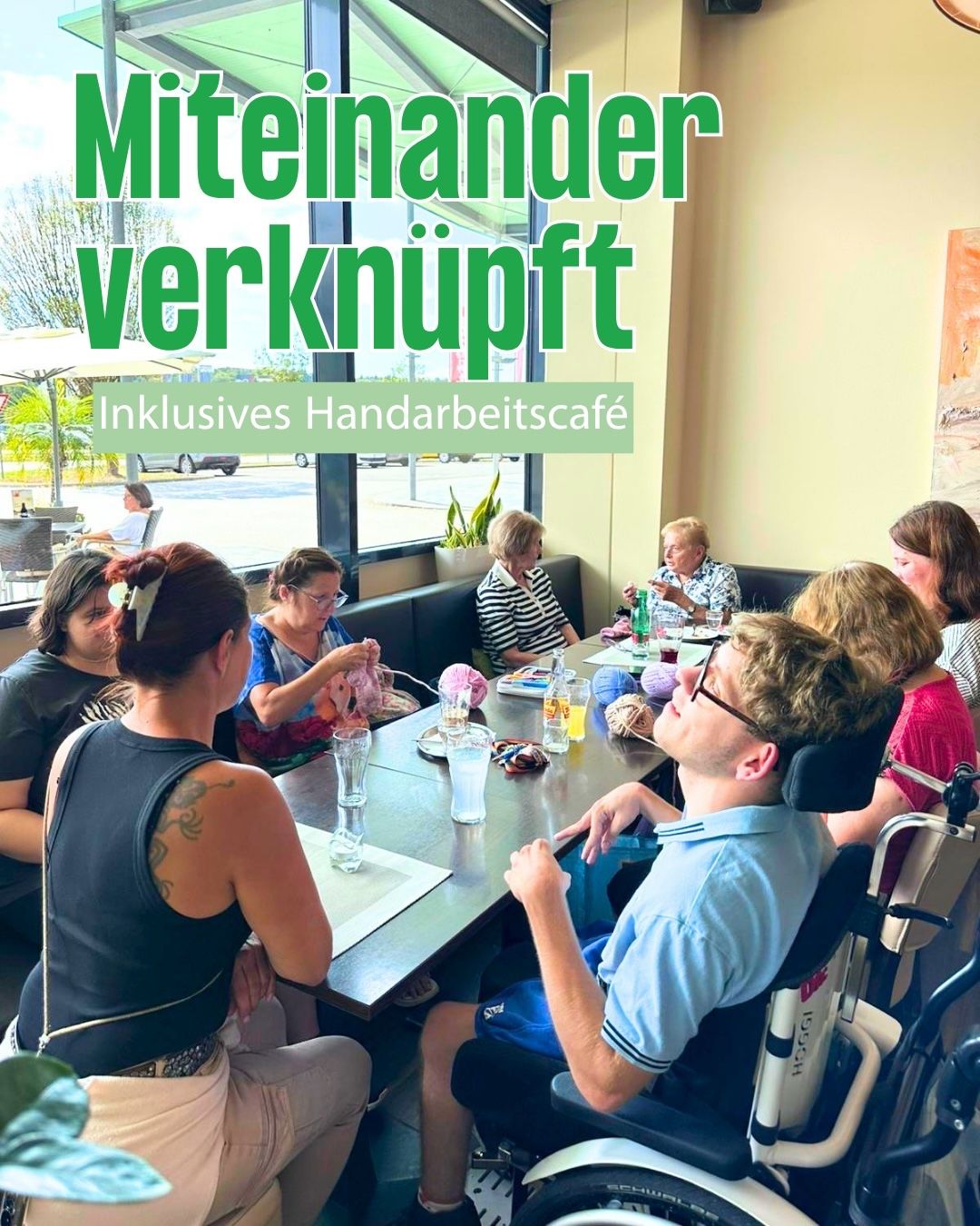 A group of people, including a man in a wheelchair, are gathered around a table in a cafe, engaged in a knitting activity. The cafe has large windows and a sign that reads 'Miteinander verknitpft'.