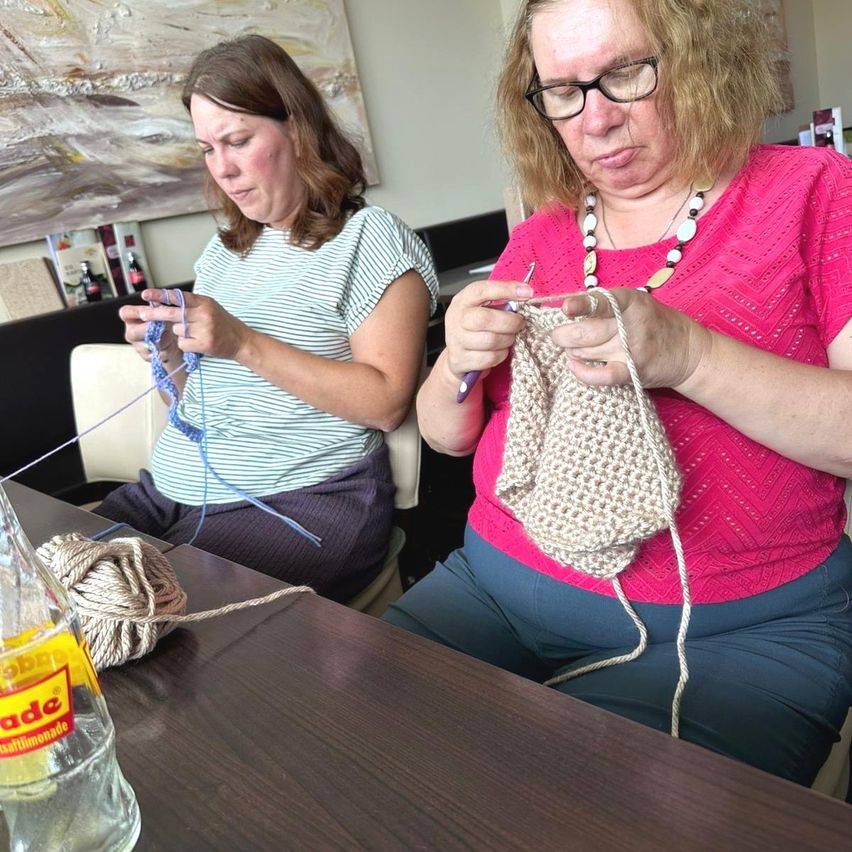 Two women are sitting at a table, knitting with yarn. One woman is holding a crochet hook and a ball of yarn. The other woman is holding a ball of yarn and knitting. A bottle of lemonade is on the table.