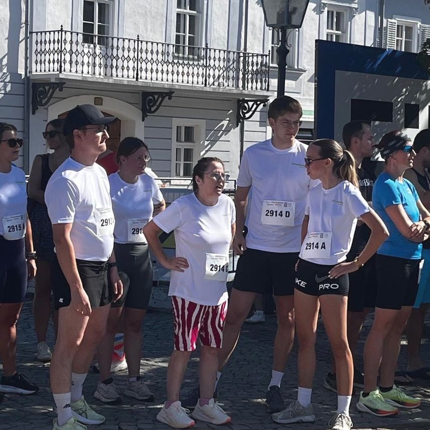 A group of people wearing white t-shirts and black shorts with numbers are standing in a line, possibly waiting for a race to start. They are in front of a building with a balcony.