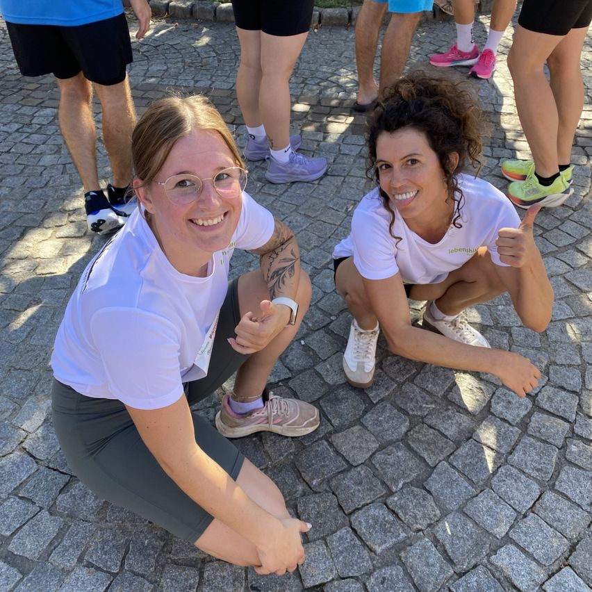 Two women in white shirts and sneakers pose for a photo on a cobblestone path, smiling and giving thumbs up.