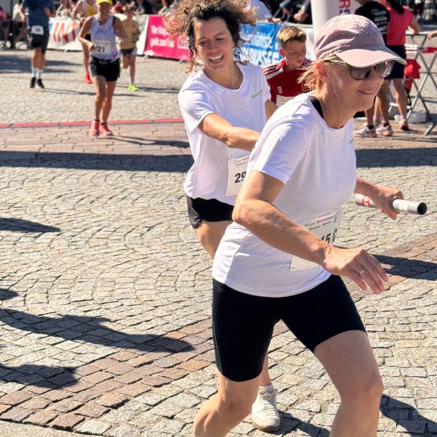 Two women in white shirts and black shorts run in a marathon. One woman wears a pink hat and glasses. They run on a cobblestone street.