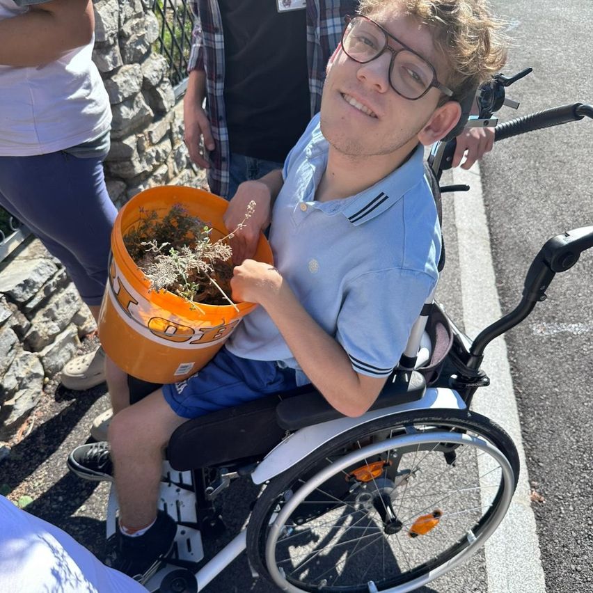 A young man in a wheelchair holds a bucket filled with plants, smiling as he poses for a photo with two people standing behind him.