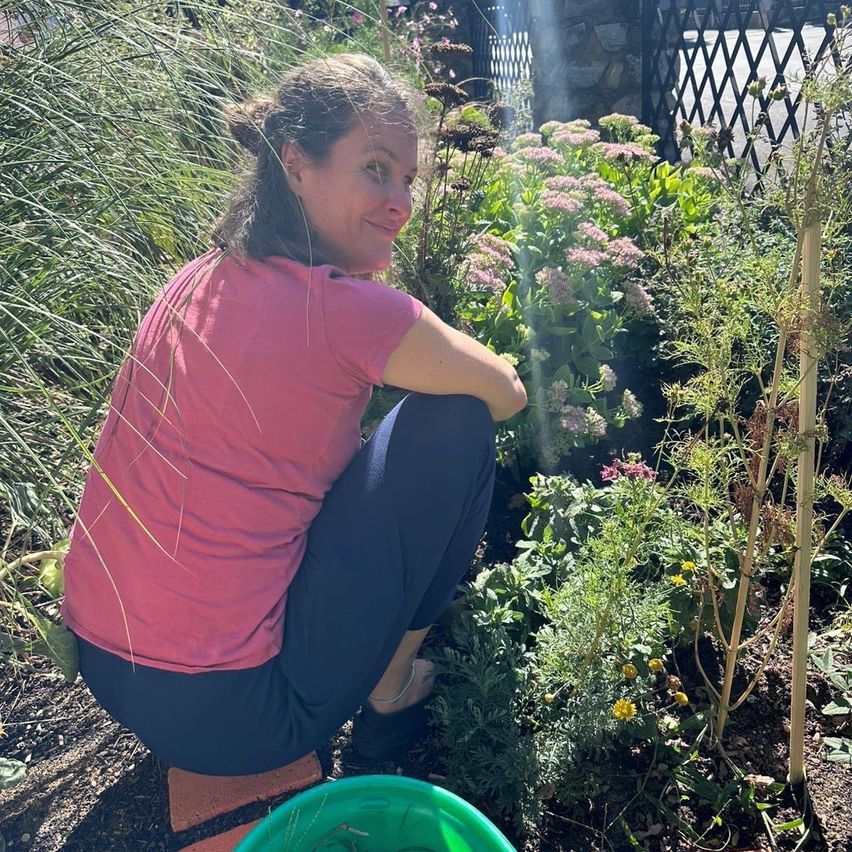 A woman in a pink shirt and blue pants is kneeling in a garden, smiling, with a green bucket nearby.
