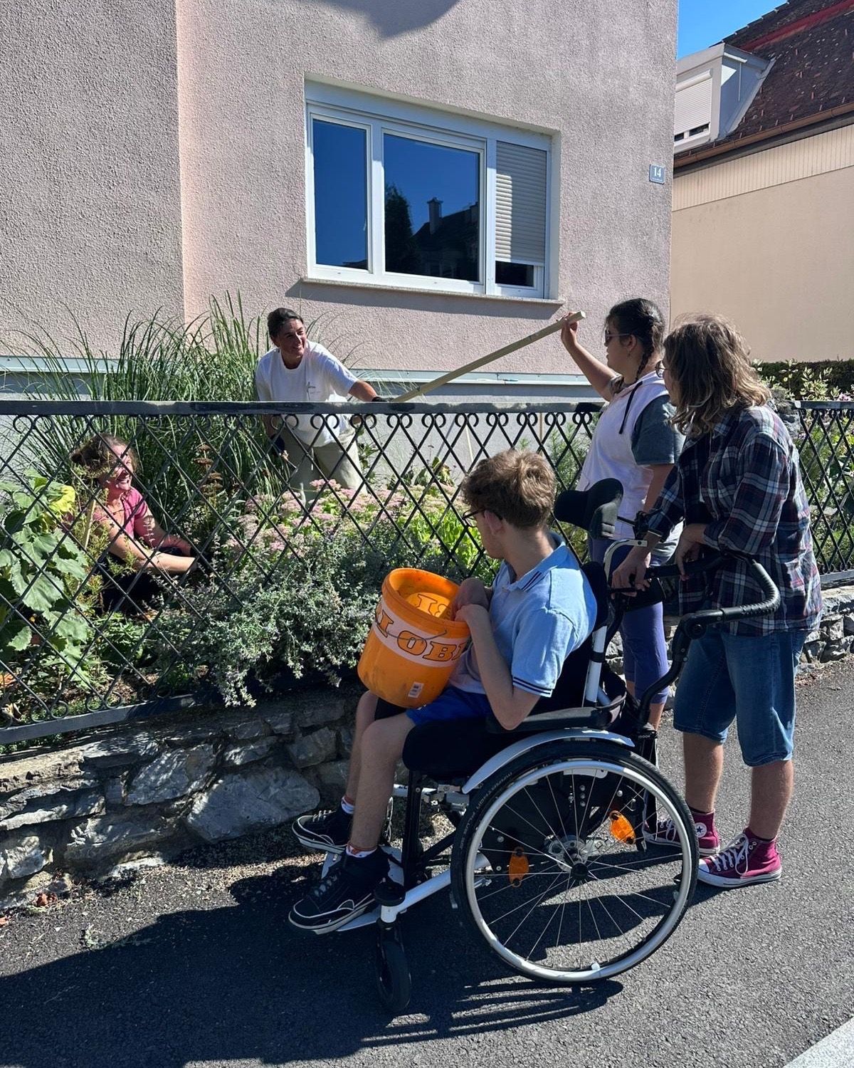 A group of people are gardening outside a house. A boy in a wheelchair holds a bucket. A woman holds a rake. Another woman crouches near plants. A man stands behind a fence.