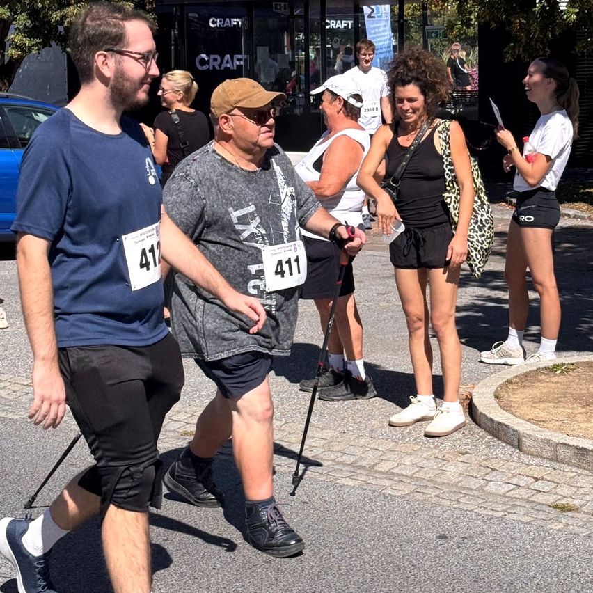 A group of people are walking on a street, possibly participating in a marathon. A man in the front is holding a cane. Behind him, a woman is holding a cup. Another woman is holding a piece of paper. There are shops on the side of the street.