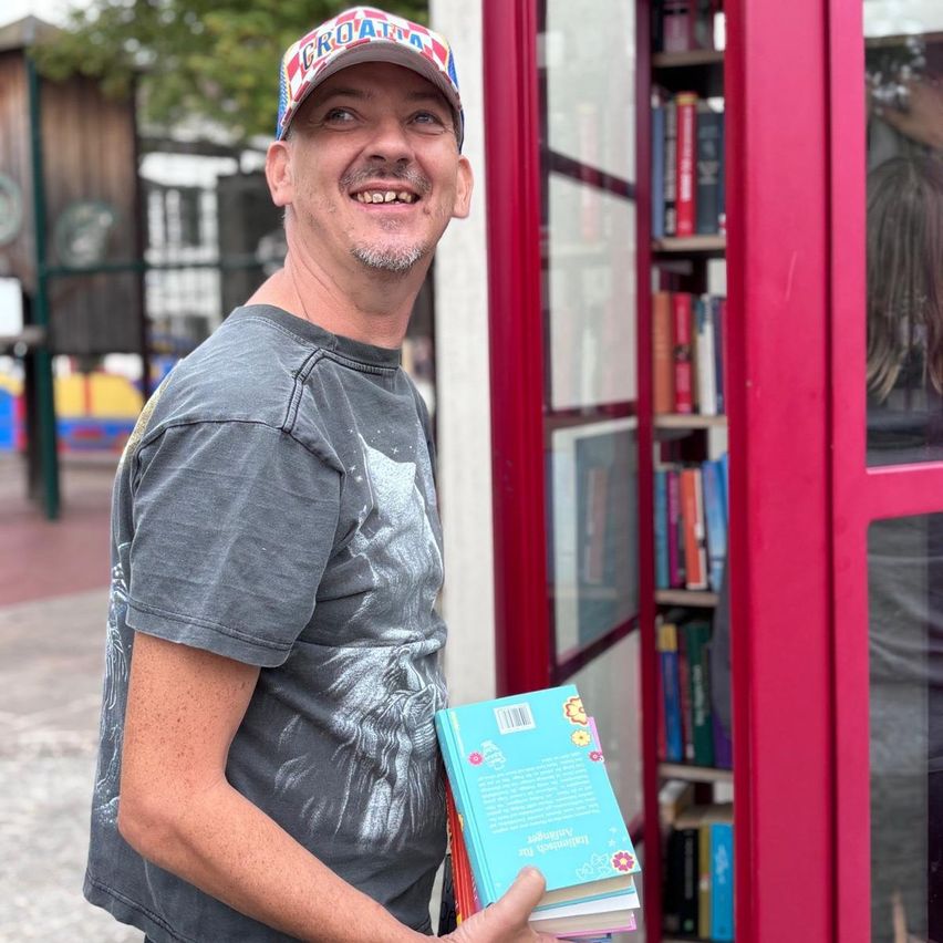 A man in a gray t-shirt and a cap is holding a book in front of a red telephone booth.