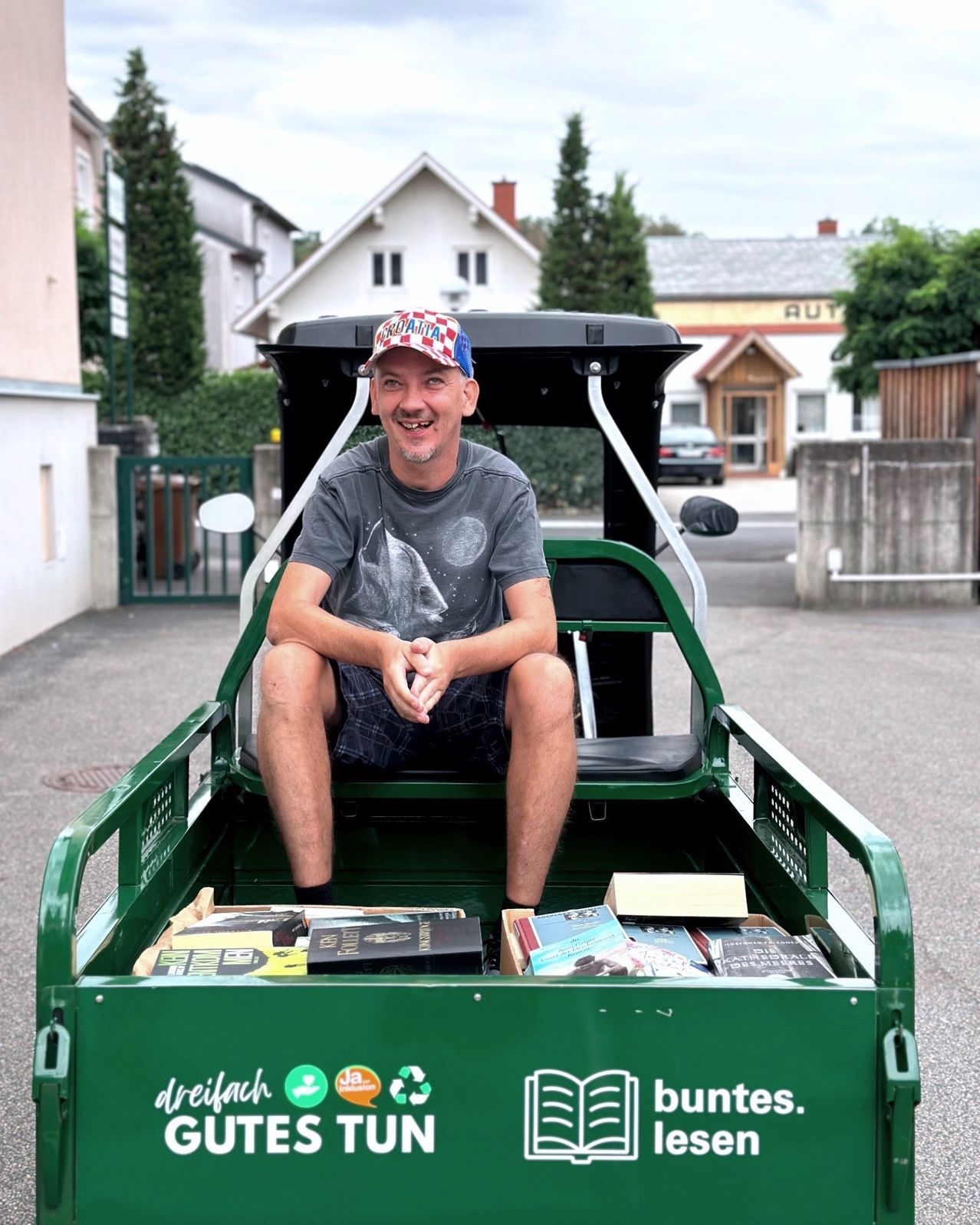 A man sits in a green cart filled with books, wearing a hat and a gray shirt. He is smiling and seems to be posing for a photo. The cart has the word 'dreifach' on it.