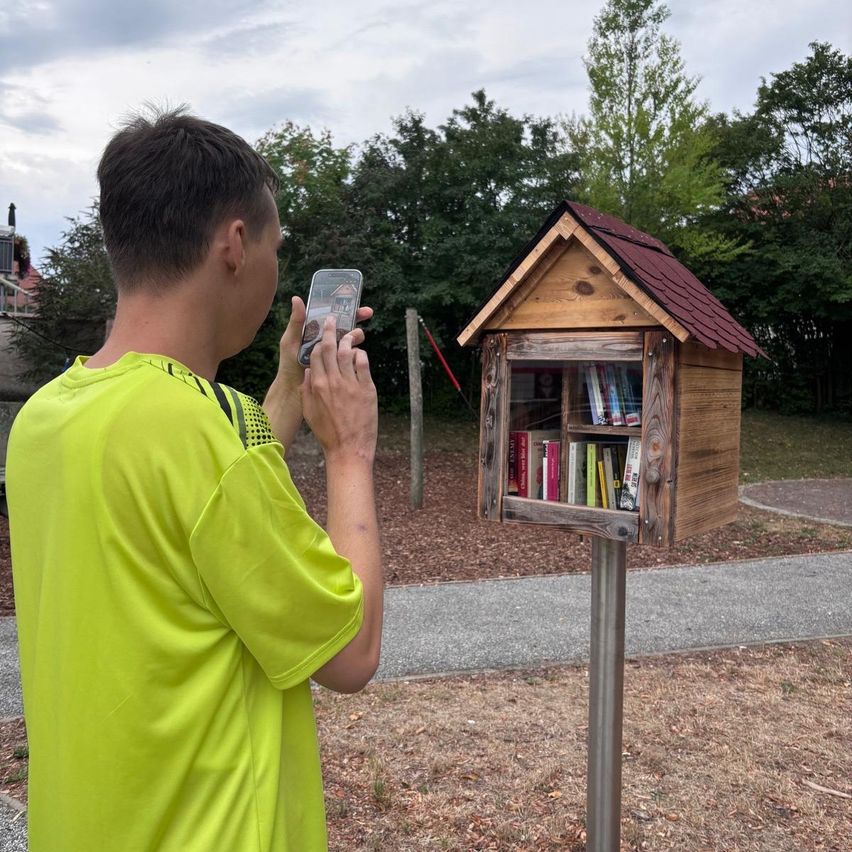 A man in a bright yellow shirt is taking a photo of a small wooden library box filled with books.