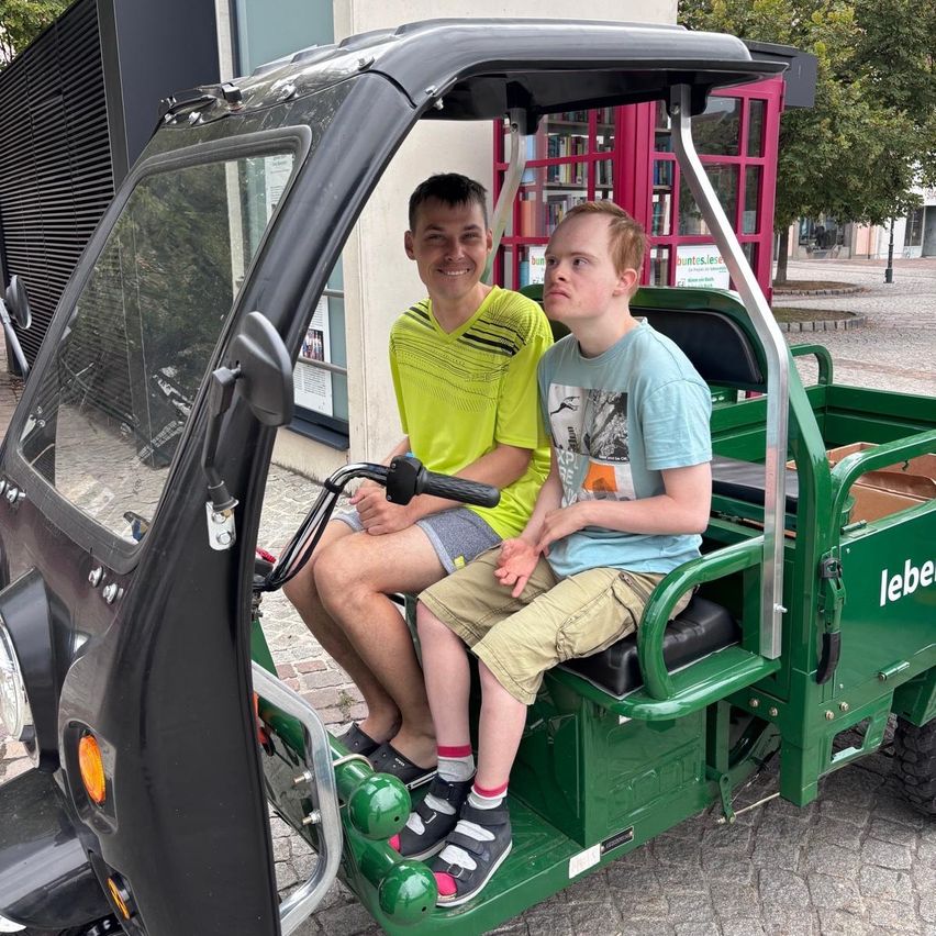 Two individuals are seated in a green electric vehicle, with one wearing a yellow shirt and the other in a blue shirt. They are both smiling and appear to be posing for a photograph.