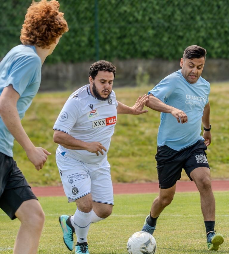 Three men play soccer on a field. One man in white looks focused. Another in blue tries to block him. A third man in blue watches from the side.