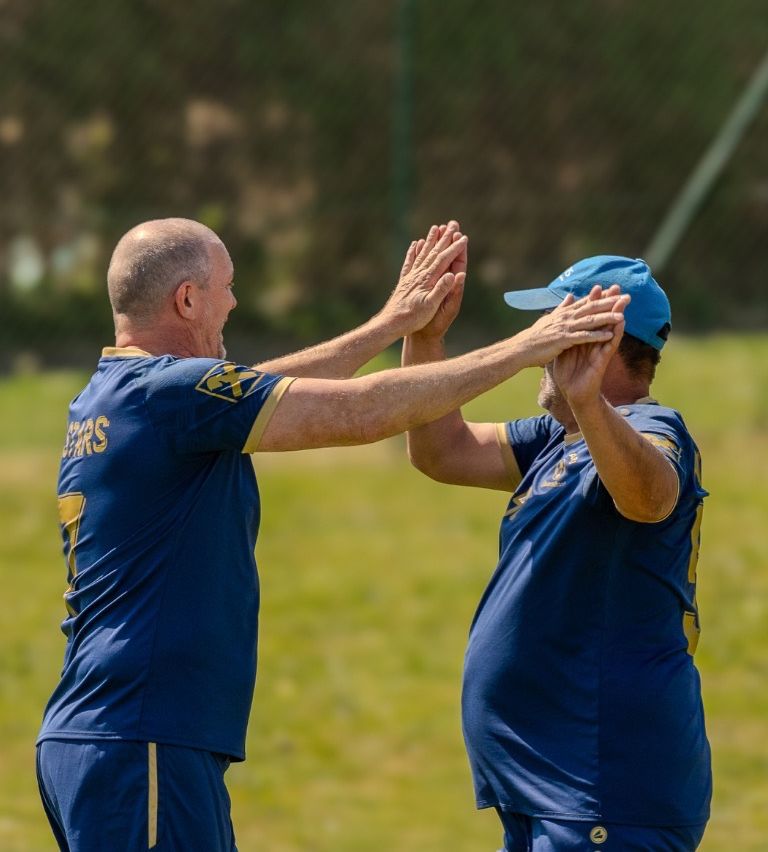 Two men wearing blue jerseys with yellow accents are clapping their hands together outside on a sunny day.