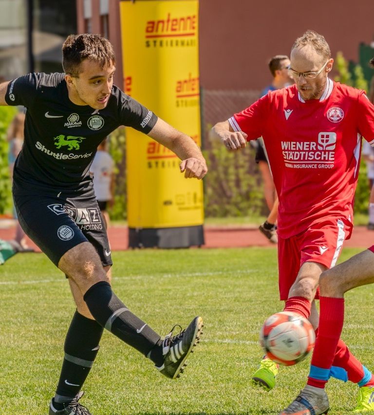 A soccer player in a black uniform is kicking the ball while another player in red tries to block. A man in glasses watches them.